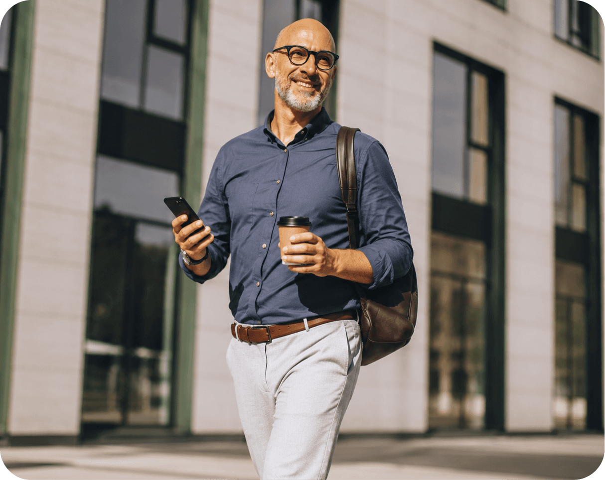 Un homme souriant, smartphone et gobelet de café à la main, marche devant un immeuble de bureaux.